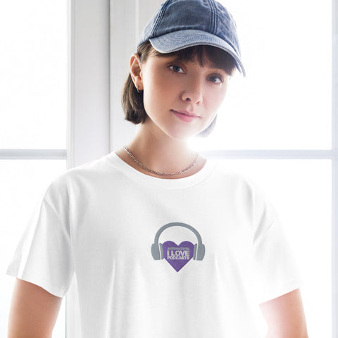 A woman wearing a Boss Uncaged Store white Affirmation I Love Podcasts Women's crop top, with headphones, showcasing her love for podcasts through a purple heart accessory.