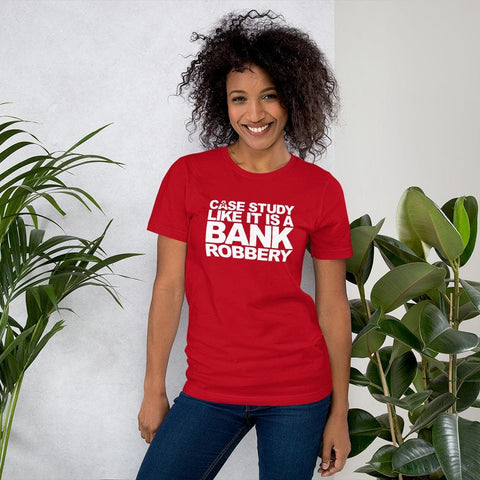 Image of A woman with curly hair wearing a red shirt that says, "CASE THE STUDY LIKE IT IS A BANK ROBBERY! GIMME THE LOOT!" by Buteke, standing between two green plants and smiling.