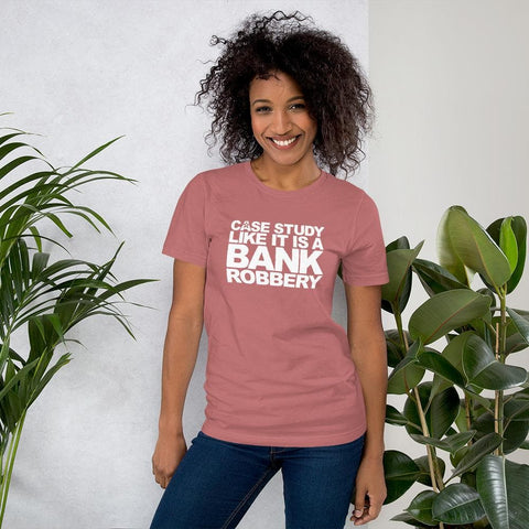 Image of A person with curly hair is smiling and wearing a pink shirt from Buteke that reads "CASE THE STUDY LIKE IT IS A BANK ROBBERY! GIMME THE LOOT!", standing next to potted plants against a light background.