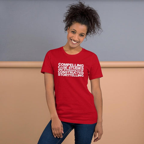 Image of A person with curly hair is smiling and wearing a red shirt from Buteke that reads, "COMPELLING CASE STUDIES ARE BYPRODUCTS OF CONSTRUCTED STORYTELLING." The background features a two-tone wall, grey on top and beige below.