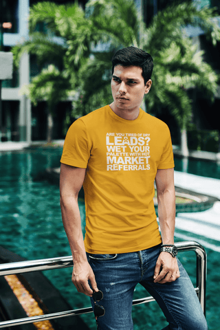 Image of A man wearing a Buteke "ARE YOU TIRED OF DRY LEADS? WET YOUR PALETTE WITH HOT MARKET REFERRALS" yellow shirt stands by a pool, leaning on a metal railing, with trees and buildings in the background, contemplating the power of referrals and networking.