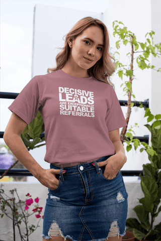 Image of A woman stands outdoors wearing a mauve t-shirt from Buteke that reads "DECISIVE LEADS ARE AN EXCEPTIONALLY SUITABLE REFERRAL" and a denim skirt. A potted plant and part of a concrete building are in the background, emphasizing the importance of generating strong leads.